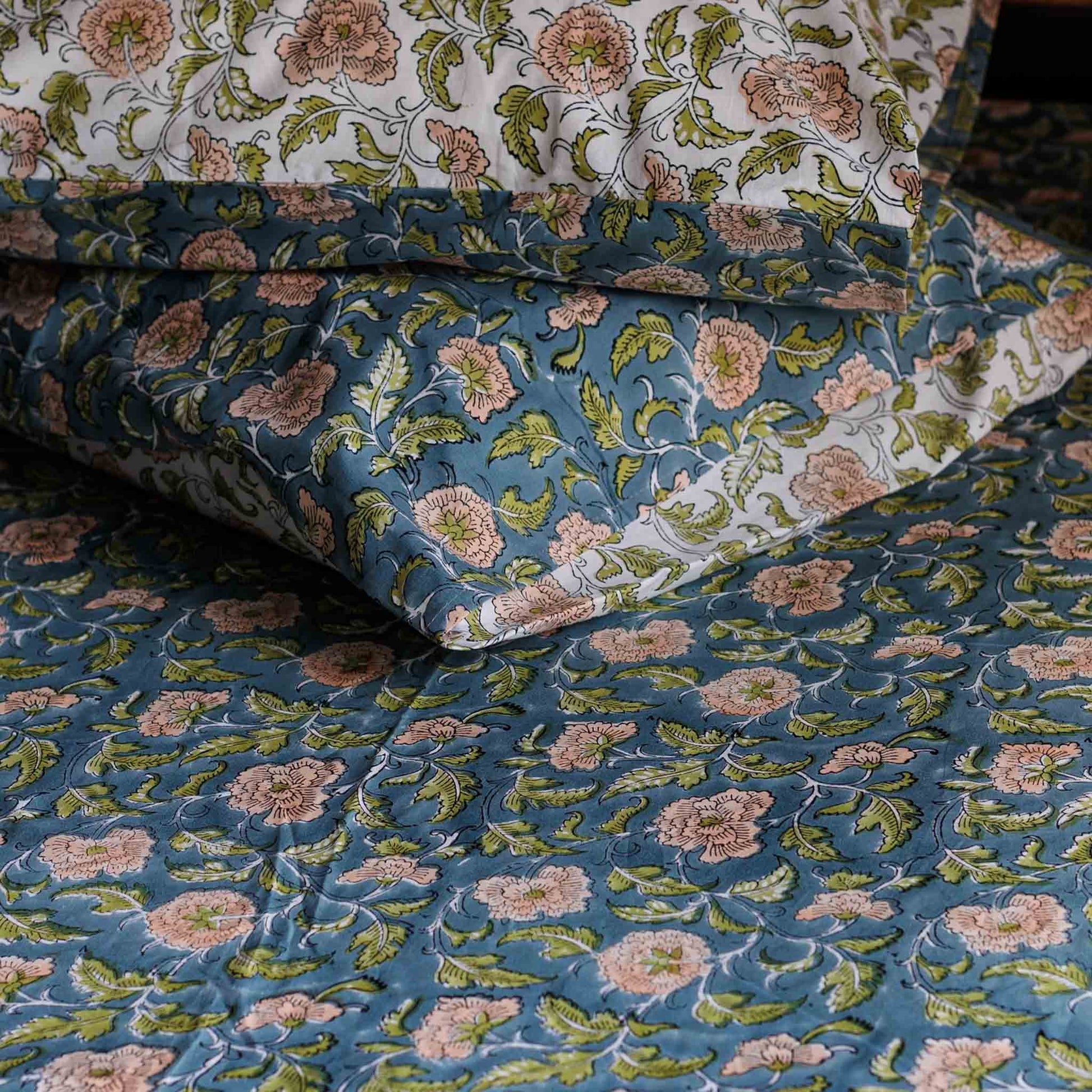 A close-up of a quilted bedding set featuring a blue base with a pink and green floral block-print pattern, including a comforter and matching pillowcases.