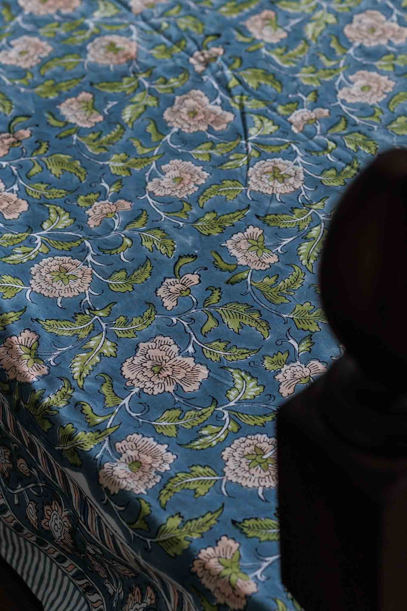 A close-up of a blue patterned textile, likely a bedspread or tablecloth, featuring a floral and leaf design in green, pink, and white. A dark wooden bed post is visible on the right side.