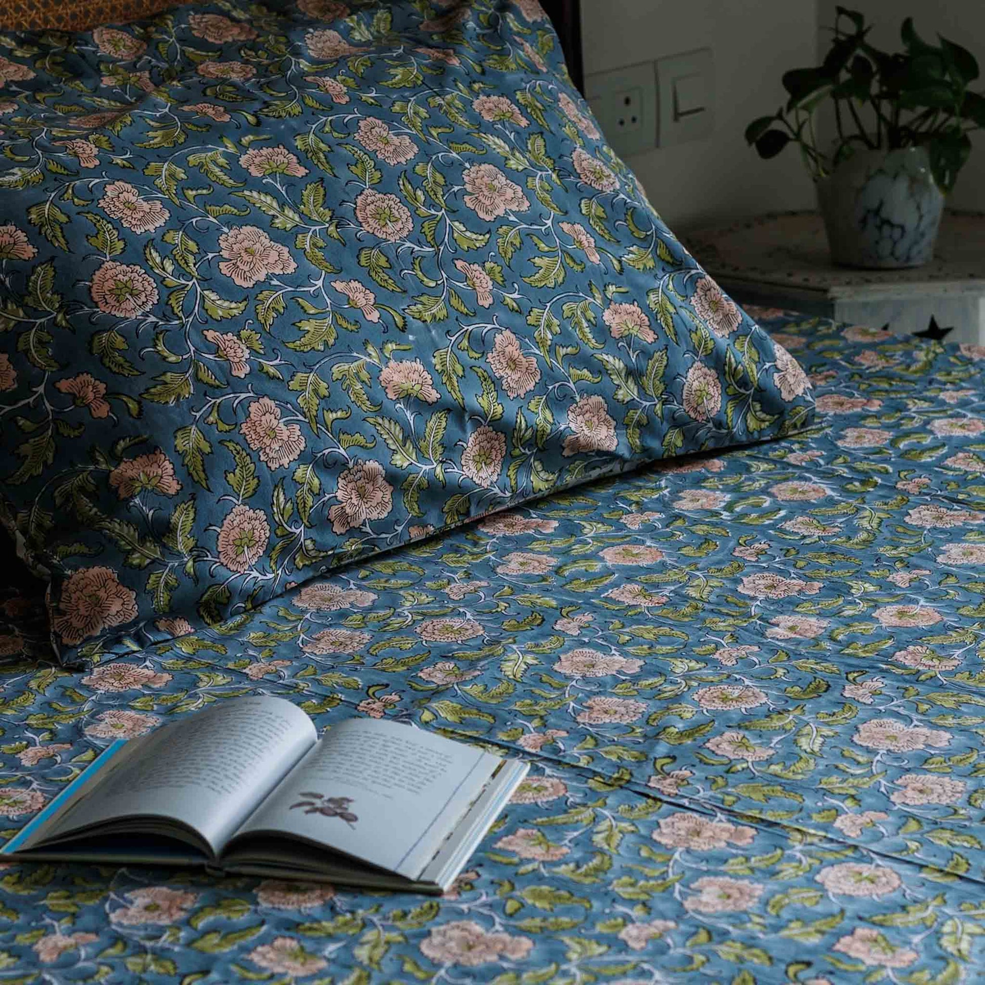 A close-up of a bed made with matching blue floral bedding, including a pillowcase and sheet, with a pattern of pink flowers and green leaves. An open book lies on the bed, and a potted plant is visible in the background near a wall outlet.