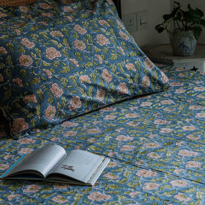 A close-up of a bed made with matching blue floral bedding, including a pillowcase and sheet, with a pattern of pink flowers and green leaves. An open book lies on the bed, and a potted plant is visible in the background near a wall outlet.