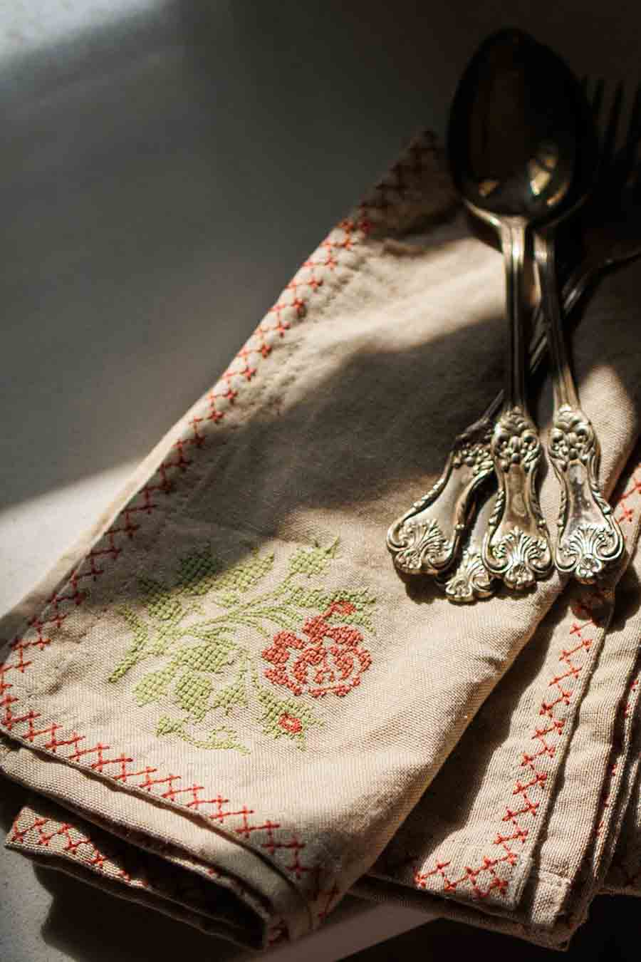 A folded cream linen napkin with red stitched border and green and red floral embroidery, styled with silver cutlery in warm, dramatic sunlight.