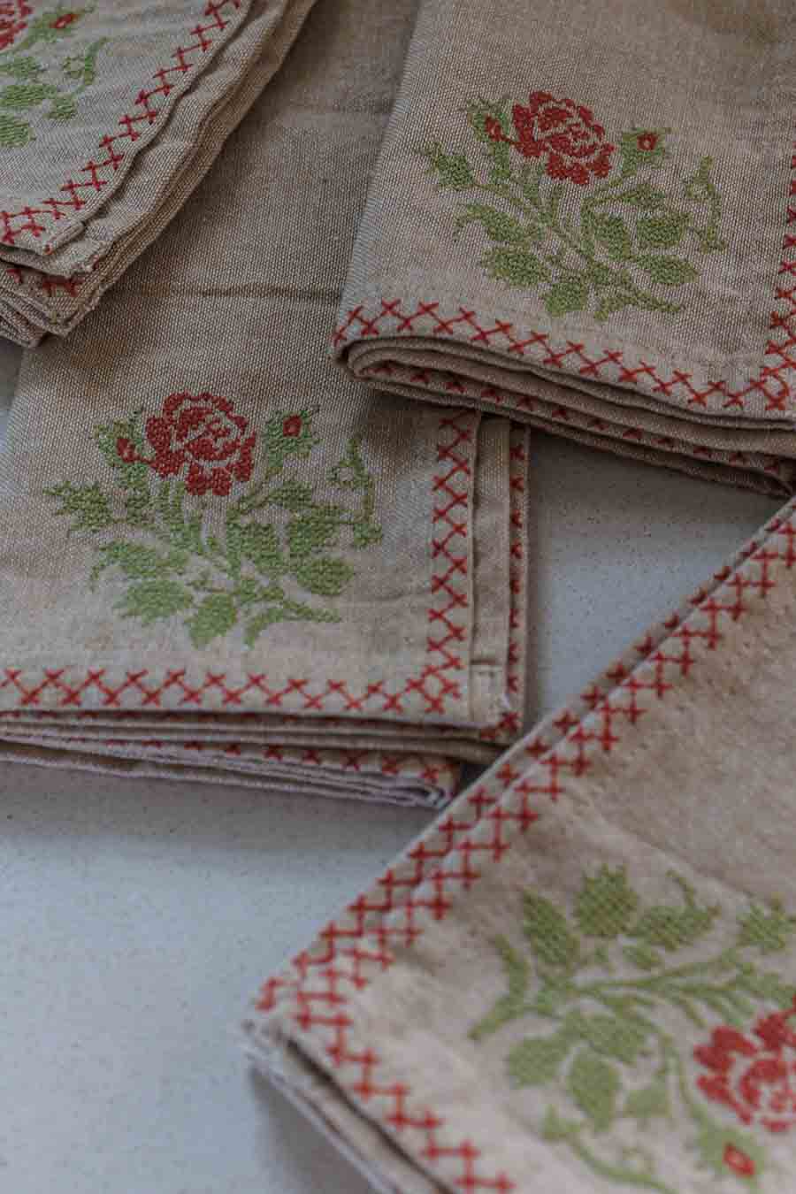 A close-up of beige napkins, featuring a red embroidered rose, green leaves, and a red stitched border on a light beige background.
