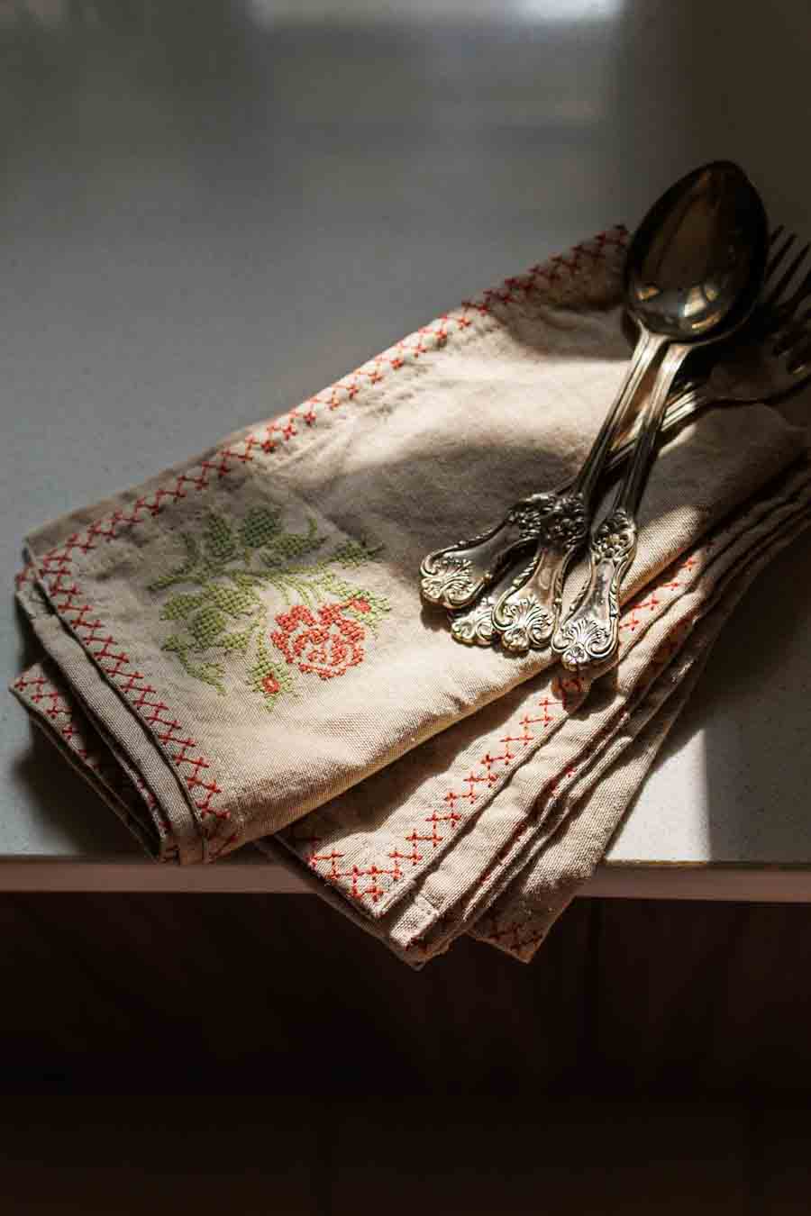 A cross stitch napkin with red stitched borders and a green floral embroidery, styled on a table with silver cutlery under dramatic, warm sunlight.