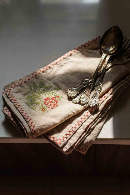 A cross stitch napkin with red stitched borders and a green floral embroidery, styled on a table with silver cutlery under dramatic, warm sunlight.