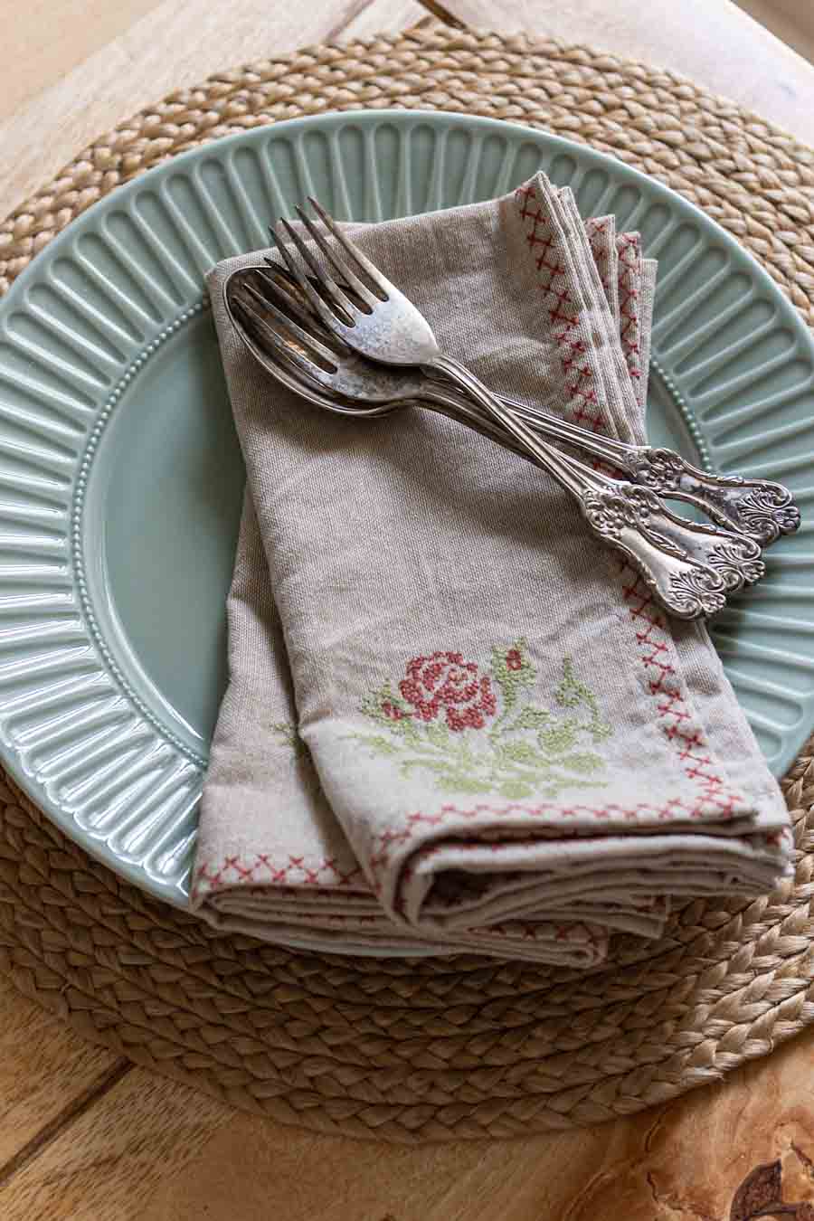 A folded beige linen napkin with a pink rose and green leaf embroidery, placed on a plate with silver forks on top.