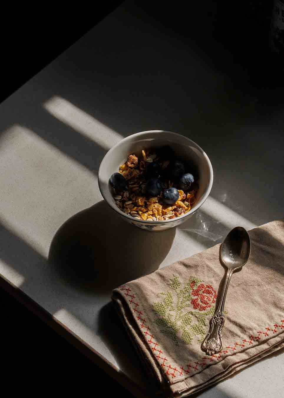 A white bowl filled with granola and blueberries sits on a tabletop in dramatic sunlight, alongside a folded beige cross stitch embroidered napkin.