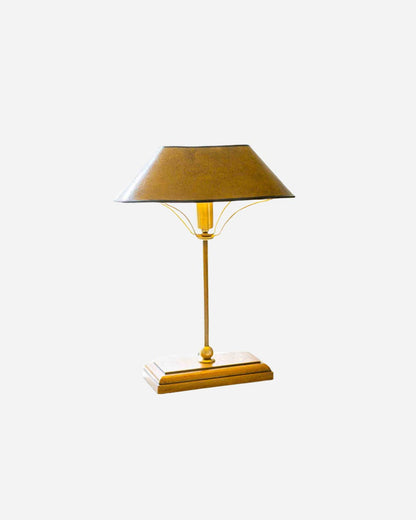 A brass table lamp with a conical shade,  a slender stem shown against a plain light gray background.