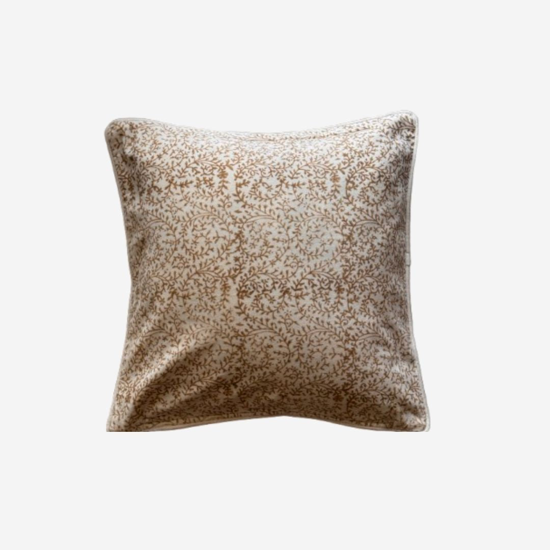A square cushion featuring block print pattern in shades of brown and beige on a light background, shown against a plain light gray studio backdrop.
