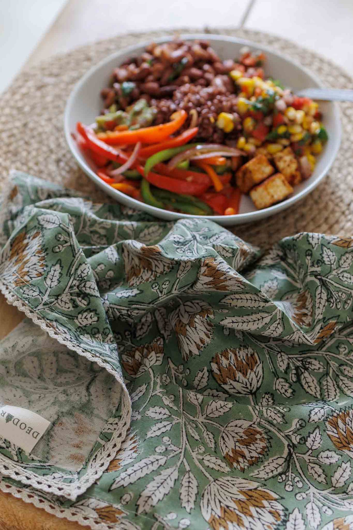 A close-up of a green floral patterned cloth napkin with lace trim, spread out in the foreground, with a bowl of colorful Mexican-style beans and rice in the background on a woven placemat.