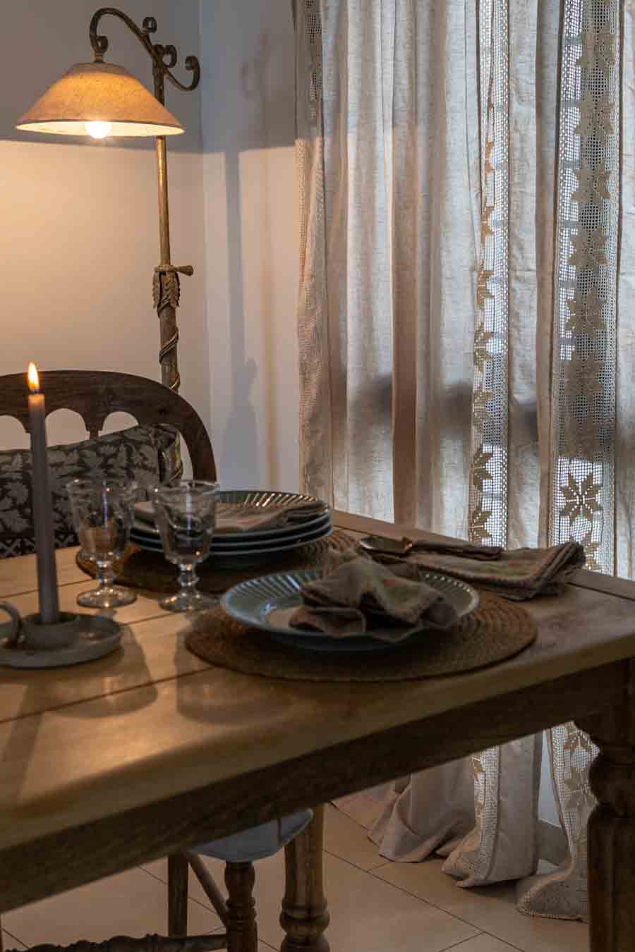 A rustic wooden dining table set with plates, folded linen napkins set against sheer lace curtains.