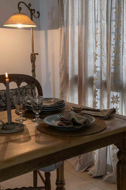 A rustic wooden dining table set with plates, folded linen napkins set against sheer lace curtains.