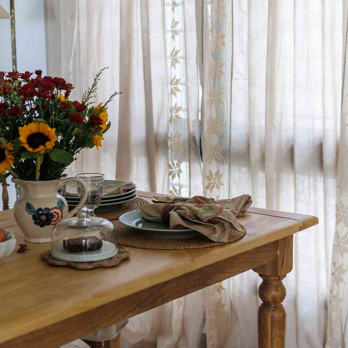 A rustic wooden dining table set with plates, a folded linen napkin, and a ceramic pitcher filled with red and yellow flowers, positioned in front of sheer lace curtains.