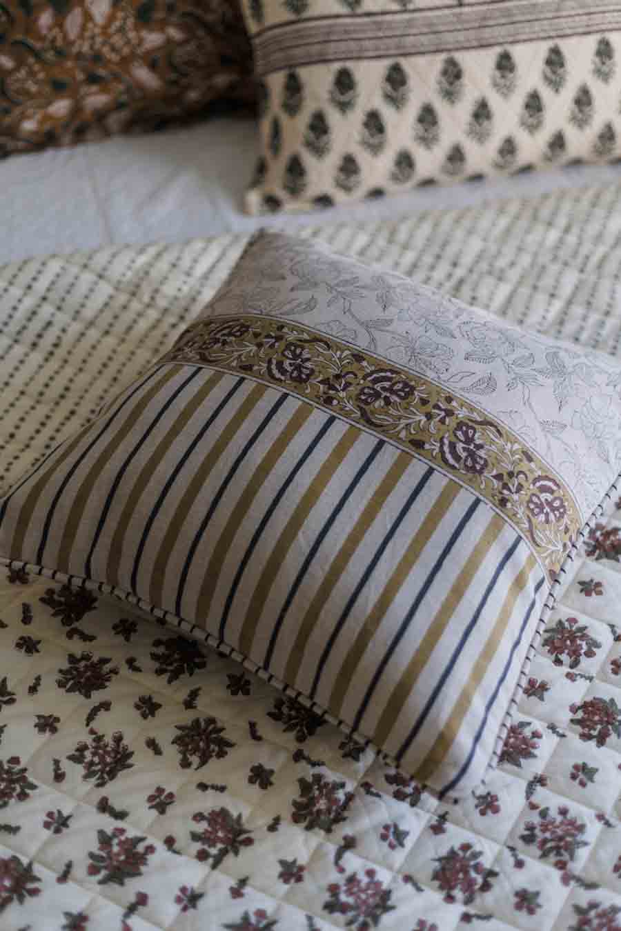A decorative cushion cover with vertical  stripes, a floral patterned top panel, and a matching embroidered border, resting on a quilted cream floral bedspread.