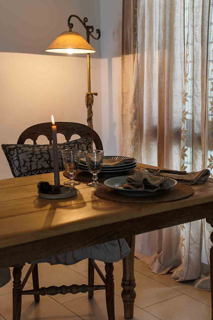 A rustic wooden dining table setting in a warmly lit room with sheer lace curtains and a floor lamp.