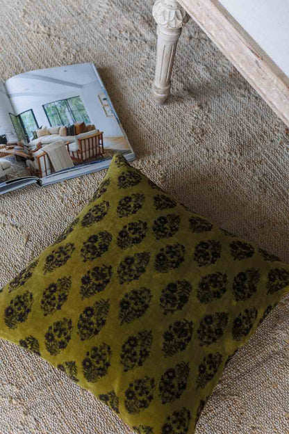 A square olive green throw pillow with a black floral block print pattern, styled on a textured beige rug next to an open magazine.