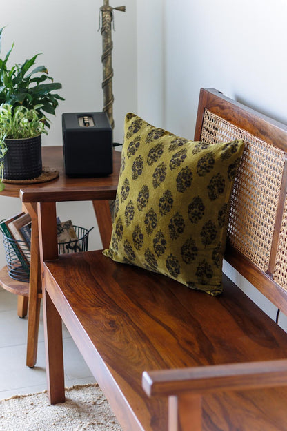 An olive green throw pillow with a black damask pattern rests on a dark wood bench with a woven cane backrest in a styled home interior.