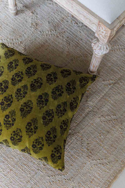 An olive green cushion with a black floral block print pattern, styled on a textured beige rug next to a distressed white wooden table leg.