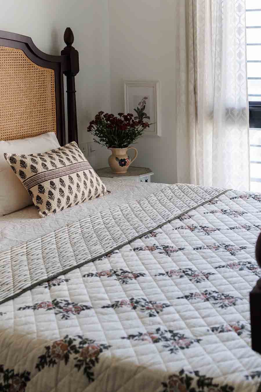 A quilted bedspread with a floral pattern and matching pillow is styled on a bed with a dark wood cane headboard in a bright, neutral bedroom.