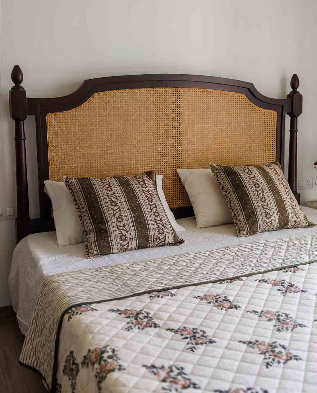 A styled bed featuring a dark wood headboard with a woven cane center panel, dressed in a cream quilted floral coverlet, striped accent pillows, and white pillows against a plain wall.