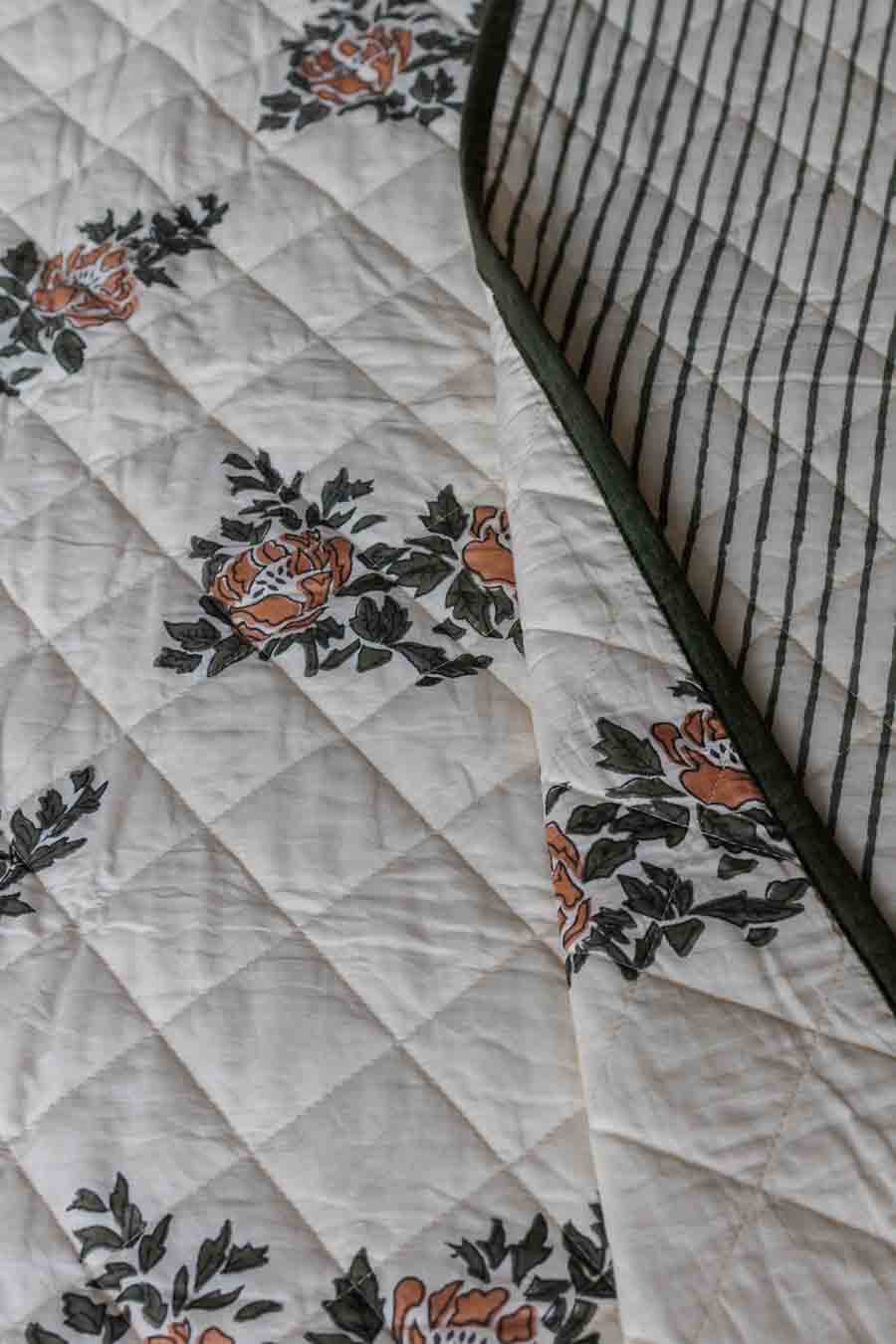Close-up of a quilted block print bedspread showing a cream background with orange floral motifs and dark green leaves, alongside a striped reverse side.