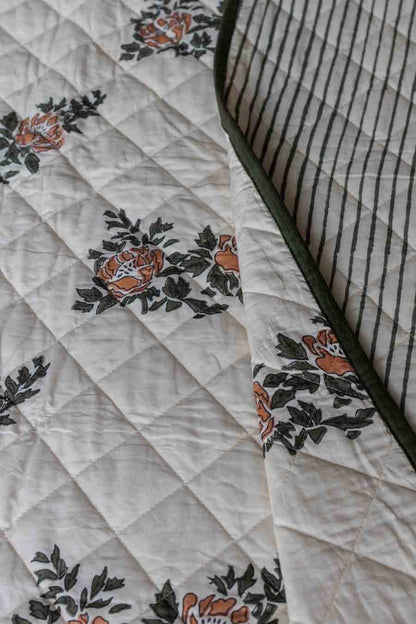 Close-up of a quilted block print bedspread showing a cream background with orange floral motifs and dark green leaves, alongside a striped reverse side.