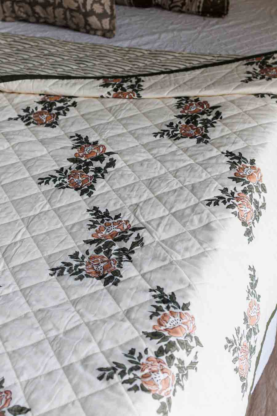 A quilted bedspread with a diamond-stitched pattern and a repeating floral print in peach, orange, and dark green on an off-white background.