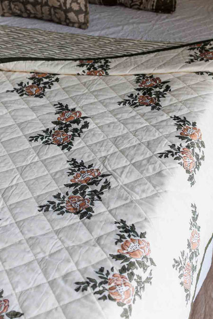 A quilted bedspread with a diamond-stitched pattern and a repeating floral print in peach, orange, and dark green on an off-white background.