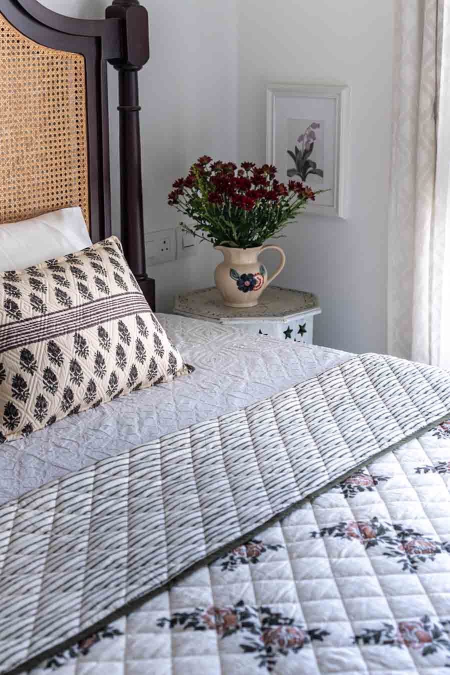 A styled bedroom scene showing a quilted bedspread with a floral pattern and striped a , alongside a patterned pillow and a dark wooden headboard.
