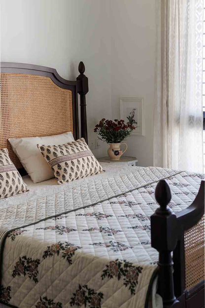 A styled bedroom scene featuring a bed with a cream quilted floral-patterned comforter, matching patterned cushions , and a dark wood frame with woven cane headboard.