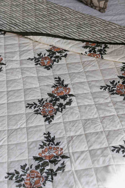 A close-up of a quilted white quilt featuring a vintage floral pattern in orange and dark green, with a striped fabric folded over at the top.