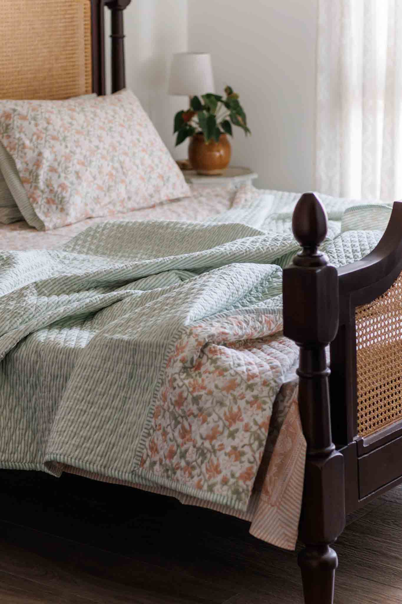 A light sage green and white striped quilted blanket with a peach and white floral patchwork section, styled on a dark wood four-poster with a woven cane headboard in a softly lit bedroom.