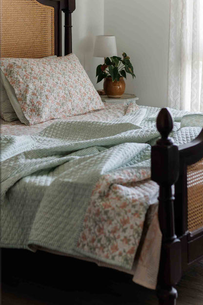 A quilted patchwork bedding set with a sage green textured section and a pink floral patterned section, styled on a bed with a dark wooden cane headboard in a softly lit bedroom.
