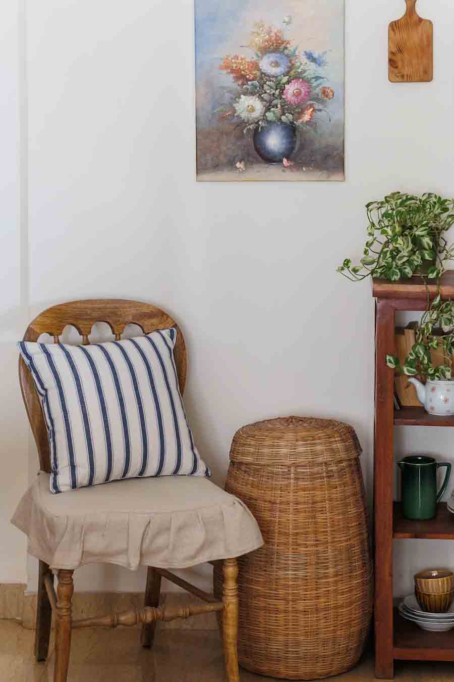 A  wooden chair with fabric seat cover and a blue and white striped cushion , positioned next to a wicker basket. set against a white wall with a floral painting, a wooden shelf with plants.