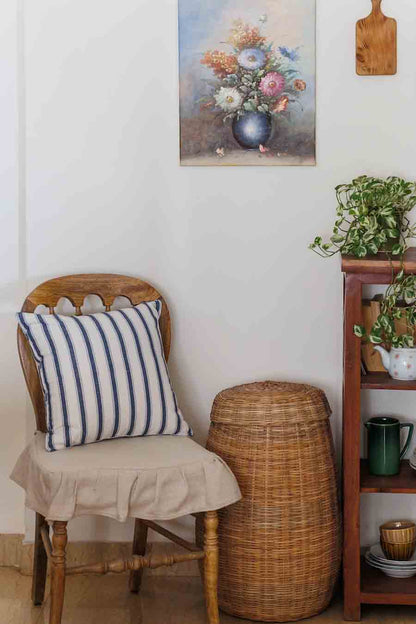 A  wooden chair with fabric seat cover and a blue and white striped cushion , positioned next to a wicker basket. set against a white wall with a floral painting, a wooden shelf with plants.