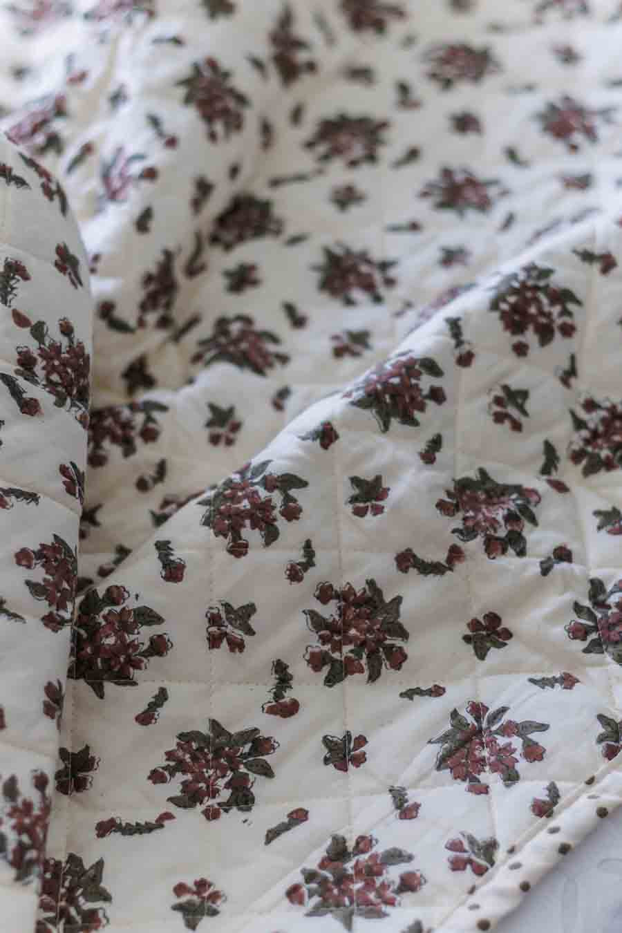 A close-up of a quilted fabric with an off-white background and a repeating floral pattern in dark green and muted red.