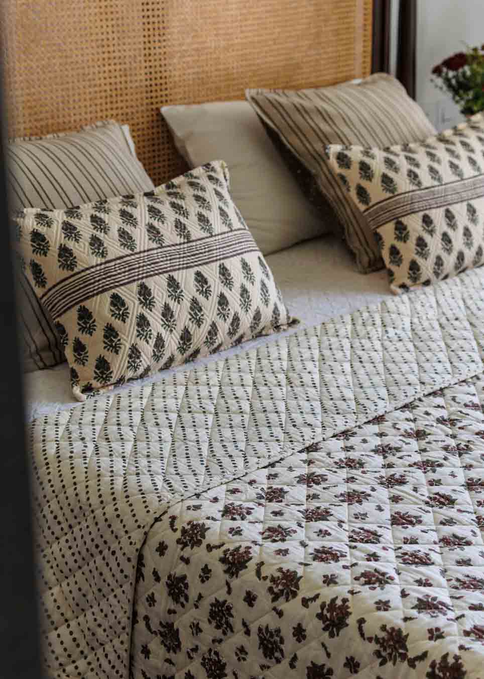 A styled bed featuring a cream quilted blanket with a dark brown floral pattern and a matching striped quilt, accented with patterned and solid pillows against a woven cane headboard.