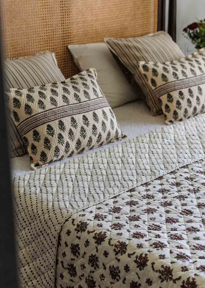 A styled bed featuring a cream quilted blanket with a dark brown floral pattern and a matching striped quilt, accented with patterned and solid pillows against a woven cane headboard.