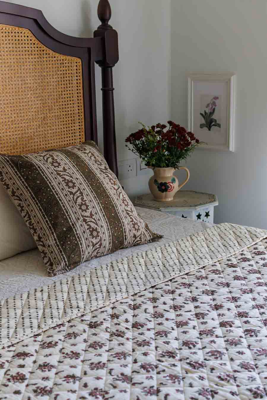 A cozy bedroom scene featuring a bed dressed in a cream quilted floral coverlet and a brown patterned accent pillow, set against a dark wood canopy headboard. A matching woven throw blanket is draped over the foot of the bed, and a small bedside table holds a ceramic pitcher of red flowers against a light gray wall.