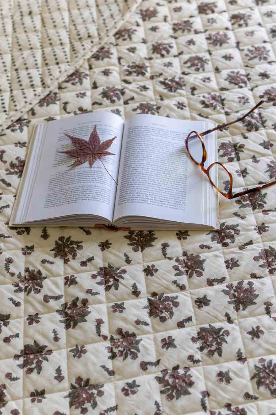 An open book with a dried red maple leaf on one page, styled on a cream quilted blanket with a brown floral pattern, with a pair of eyeglasses resting on the right page.