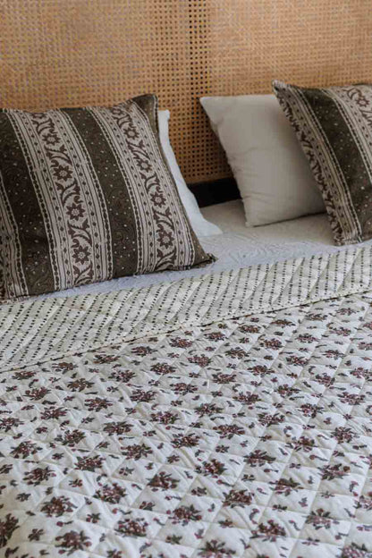 A close-up of a bed styled with a cream quilted coverlet featuring a brown floral pattern, matching patterned pillows, and a woven rattan headboard.