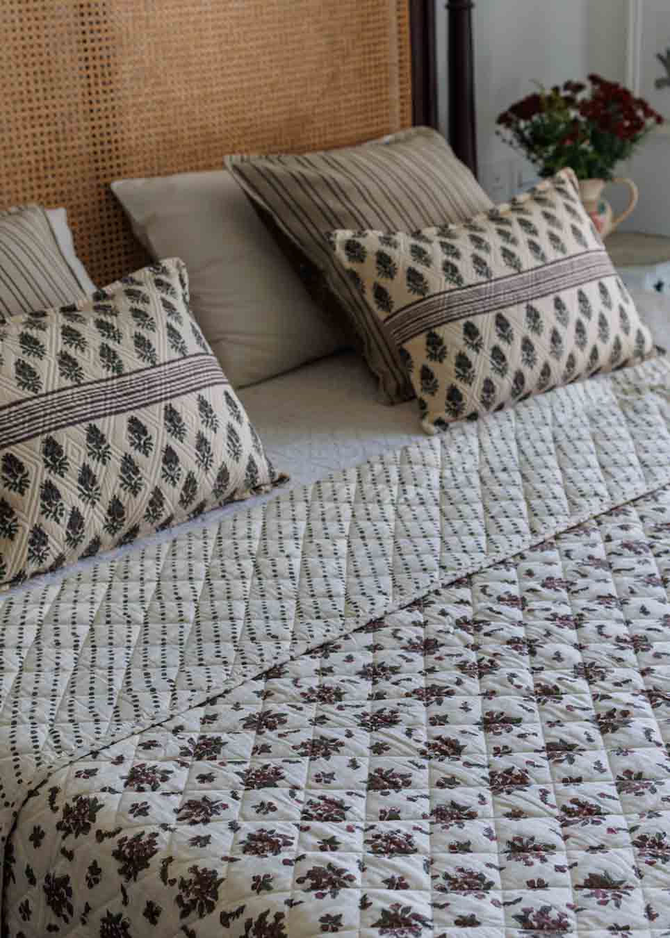 A styled bed featuring a quilted cream bedspread with a brown floral pattern, matching patterned pillows, and a woven cane headboard in a bedroom setting.