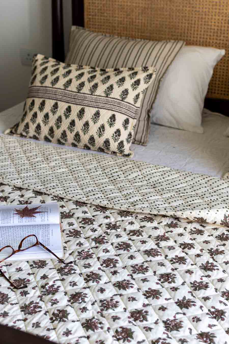A cozy bed setup featuring a cream-colored quilted blanket with a brown floral pattern, a matching patterned accent pillow, styled with an open book and glasses.
