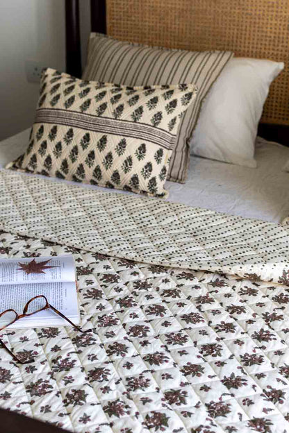 A cozy bed setup featuring a cream-colored quilted blanket with a brown floral pattern, a matching patterned accent pillow, styled with an open book and glasses.