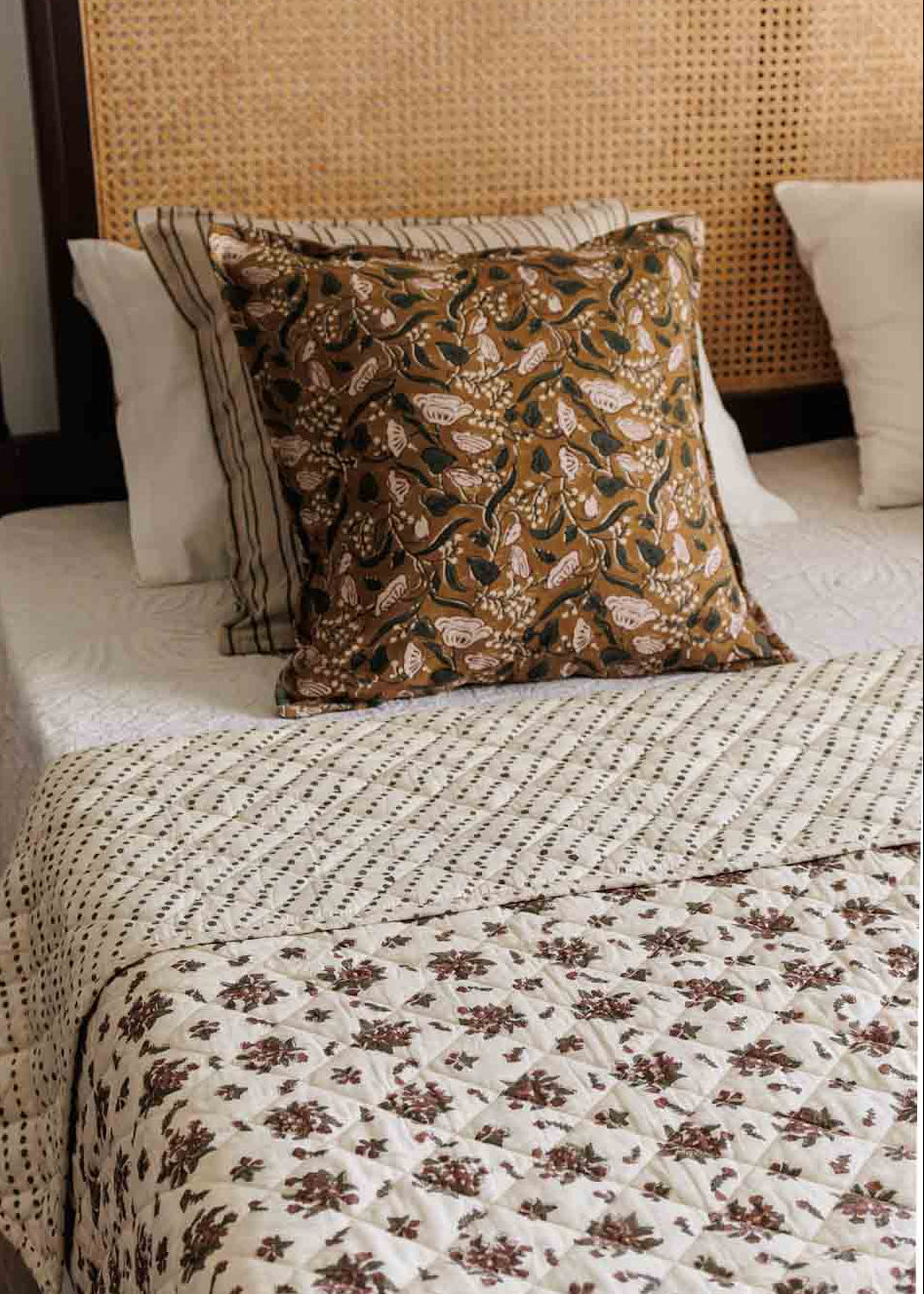 A styled bedroom showing a bed with a cream quilted coverlet featuring a brown floral pattern and two decorative cushion covers