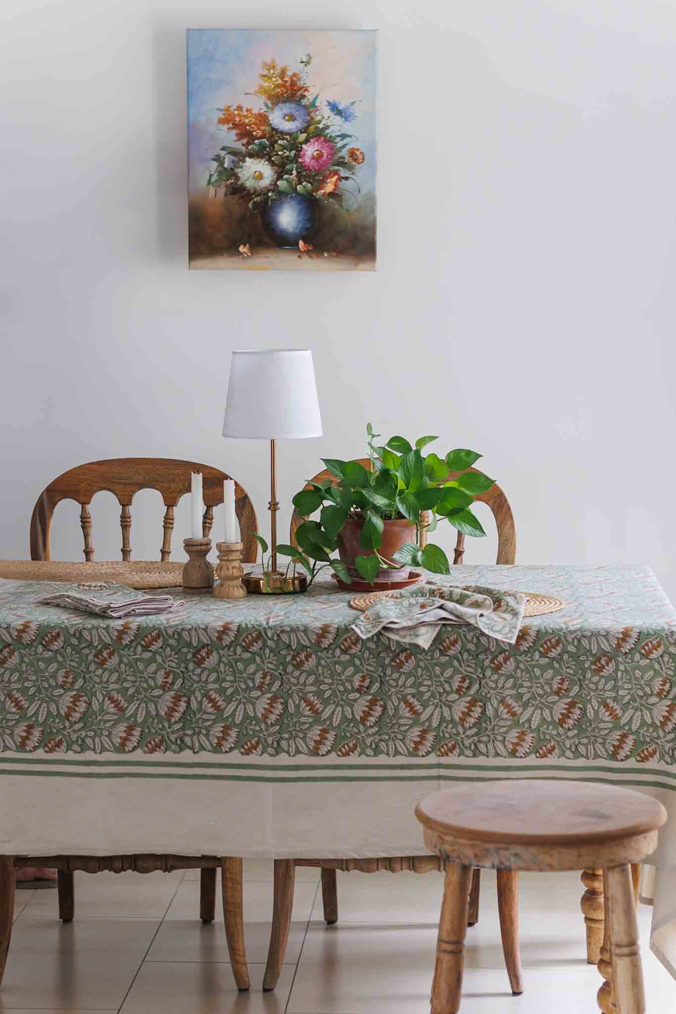 A framed floral painting hangs on a white wall above a dining table set with a floral patterned block print tablecloth, wooden chairs, a lamp, and a potted green plant.