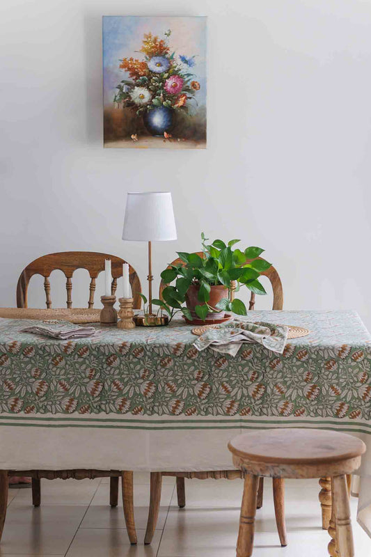 A framed floral painting hangs on a white wall above a dining table set with a floral patterned block print tablecloth, wooden chairs, a lamp, and a potted green plant.