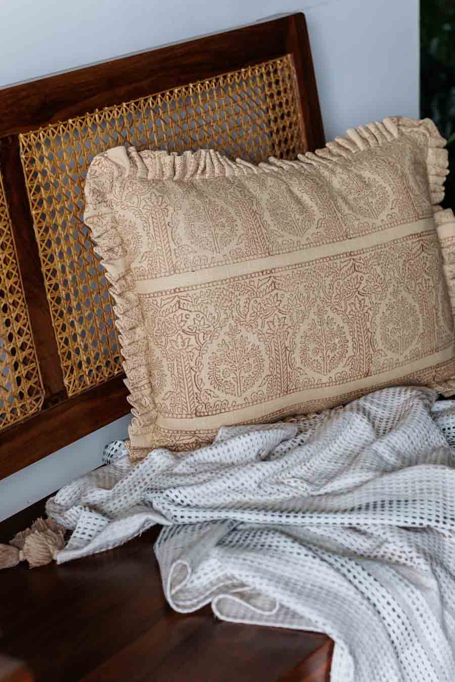 Rust block print cushion with ruffled edges, resting on a white textured blanket in front of a dark wood cane-back chair.