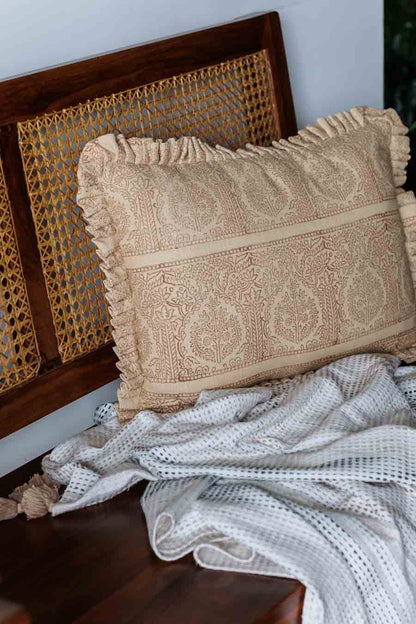 Rust block print cushion with ruffled edges, resting on a white textured blanket in front of a dark wood cane-back chair.
