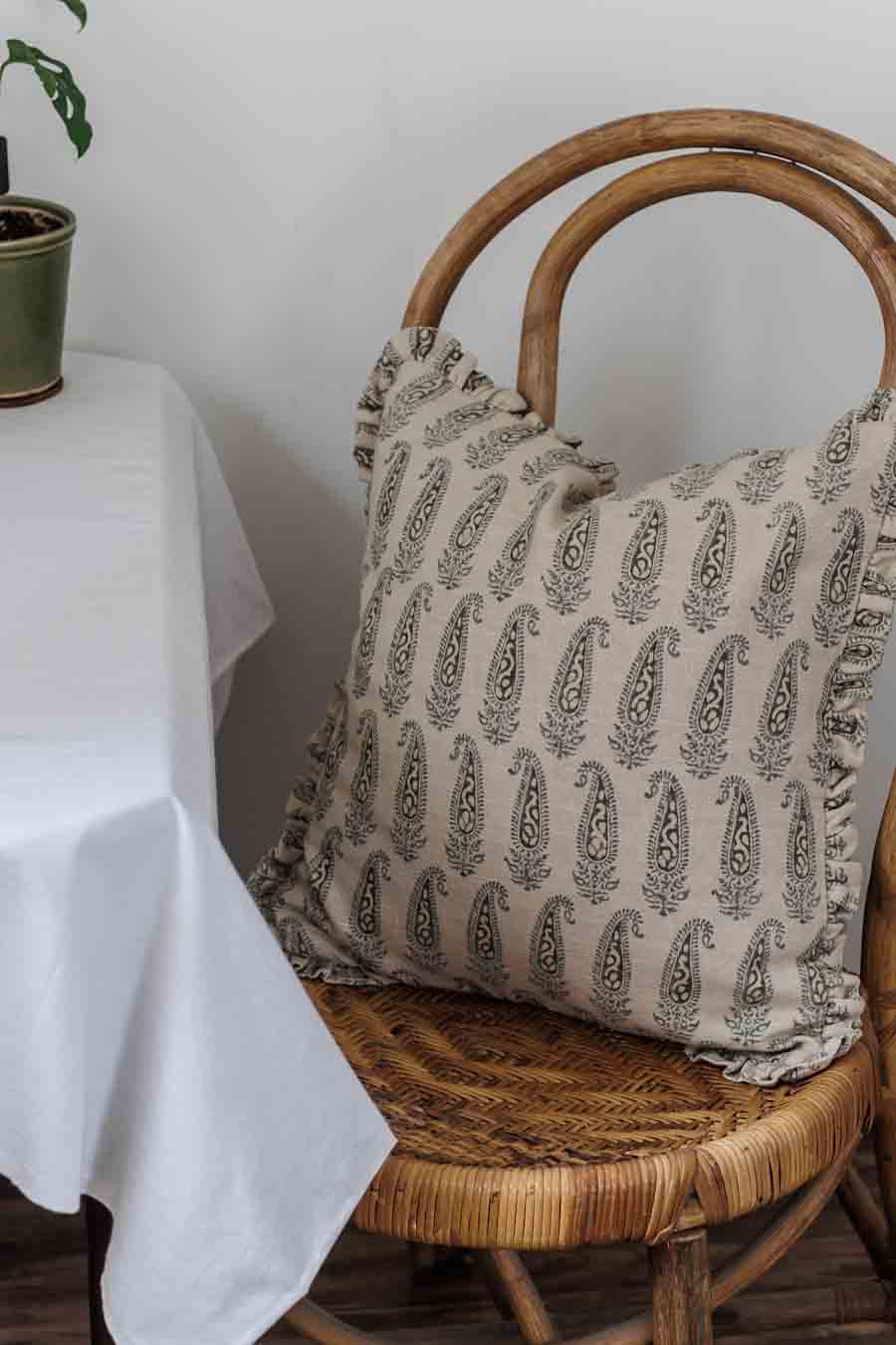 An 18 inch cushion cover with a paisley pattern in shades of green, featuring ruffles is placed on a wicker chair.