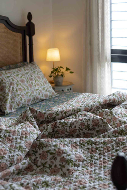 A close-up of a quilted bedding set with a pink and green floral pattern on a white background, styled on a bed in a cozy bedroom with a lit bedside lamp and window blinds.